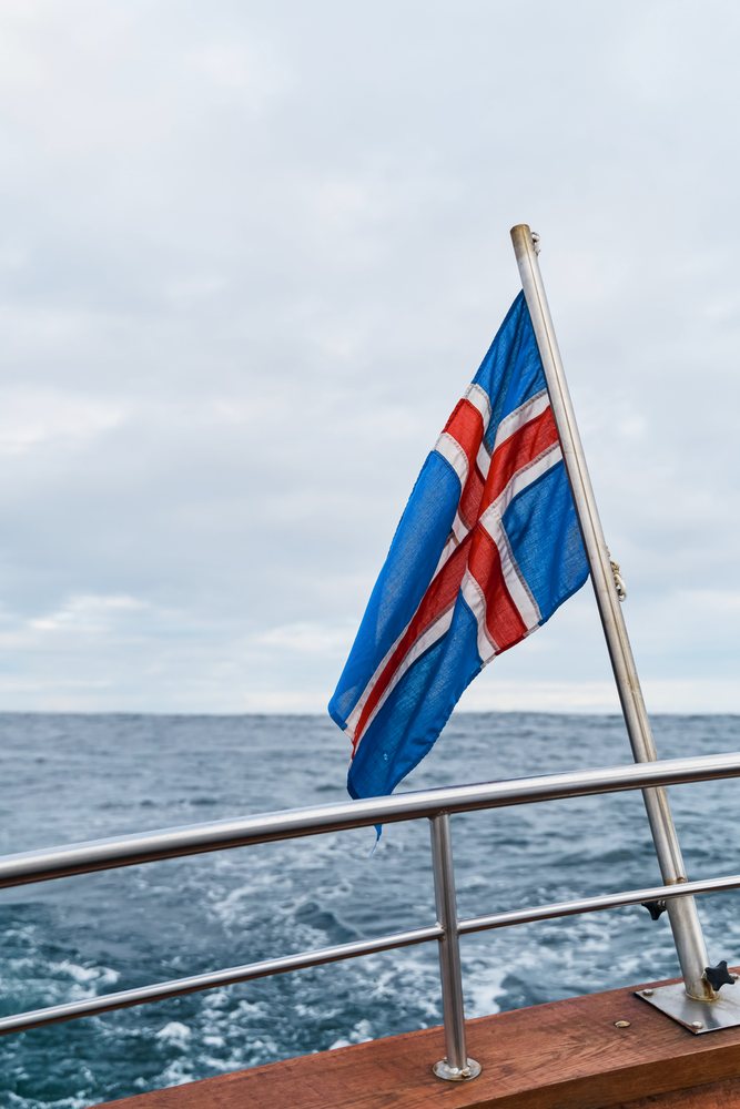 A flag with a blue background, white stripes, and a red cross fluttering on a metal pole attached to a boat railing against the backdrop of the ocean and a cloudy sky, Iceland.