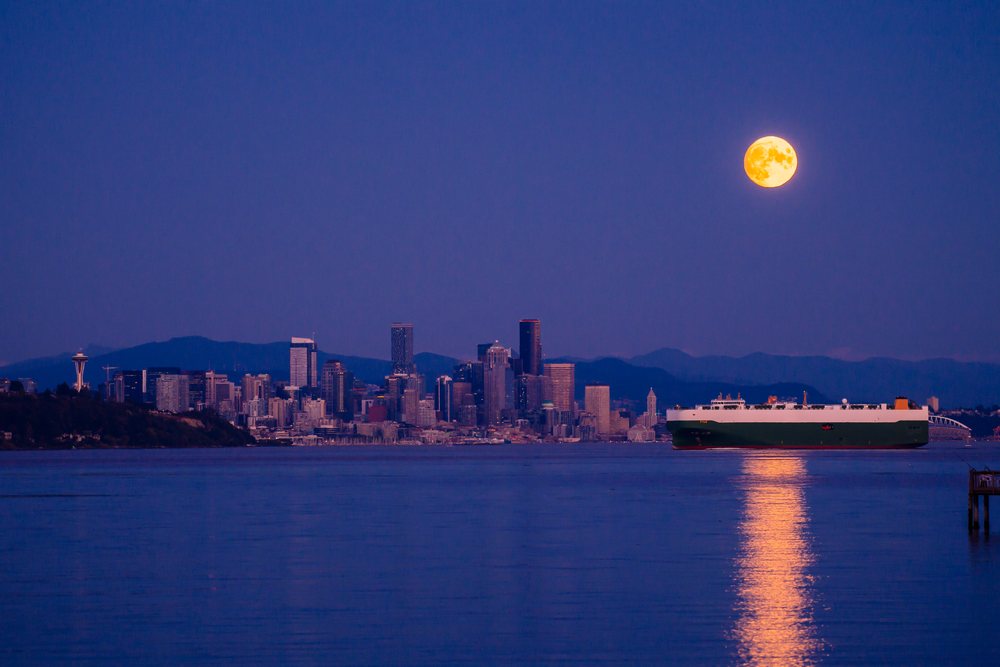 Full moon shines over Seattle skyline with cargo ship reflecting on water at night, USA.