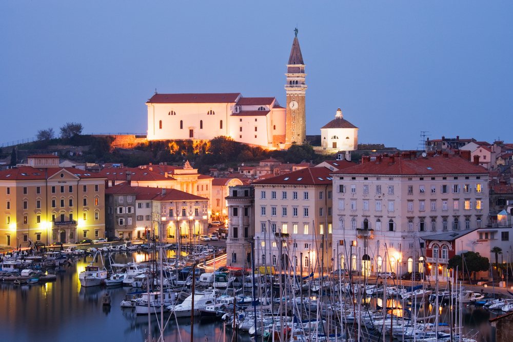 Dusk view of the Town of Piran showing illuminated buildings, a church with clock tower, boats docked in the marina, and calm water reflections, Slovenia.