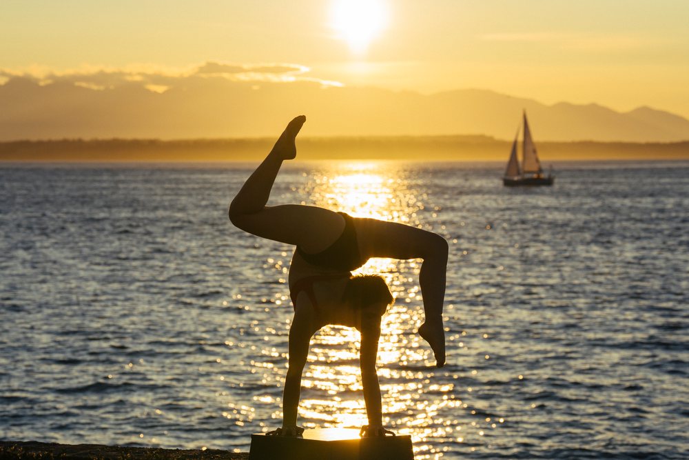 Person performs a handstand on a raised surface by the waterfront at sunset with a sailboat and mountains visible in the background, USA.