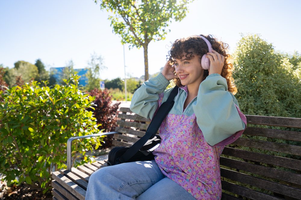 Woman with curly hair wearing floral patterned jacket and jeans sits smiling on a wooden bench in a sunny urban park, Spain.