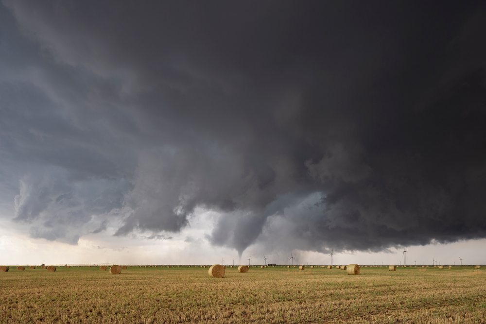 Tornado funnel cloud touches down in a flat rural field with round hay bales and wind turbines under dark storm clouds, Lingo, New Mexico, USA.