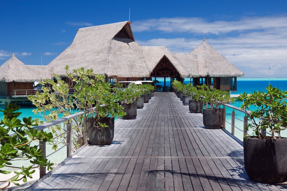 Thatched roof overwater bungalows connected by a wooden pier with potted plants over turquoise Pacific Ocean water, Tahiti, French Polynesia.
