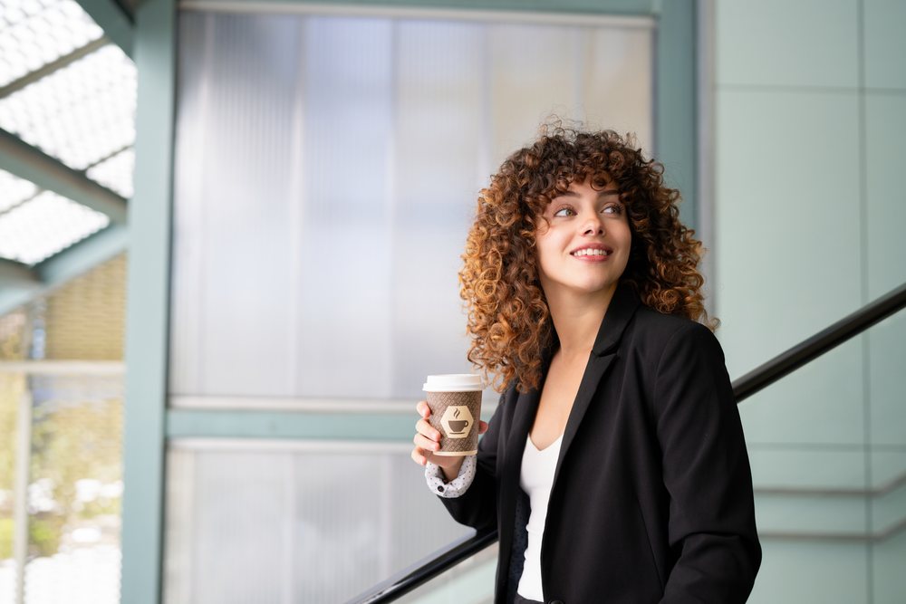 Woman with curly hair holding a disposable coffee cup stands smiling on an escalator in a modern urban setting, Spain.