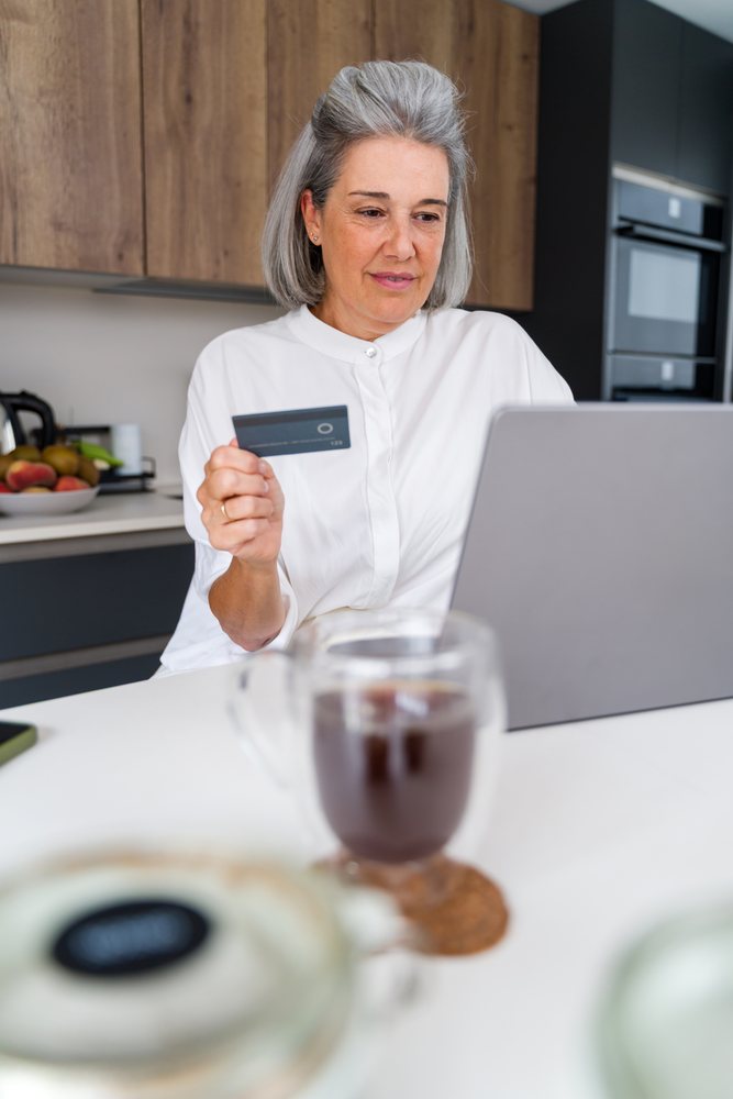 Senior woman with gray hair uses a credit card and laptop to make an online purchase in a modern kitchen, Spain.