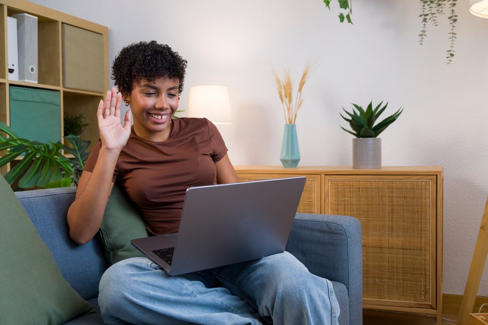 Woman with curly hair sitting on a blue sofa waving during a video call on her laptop at home.