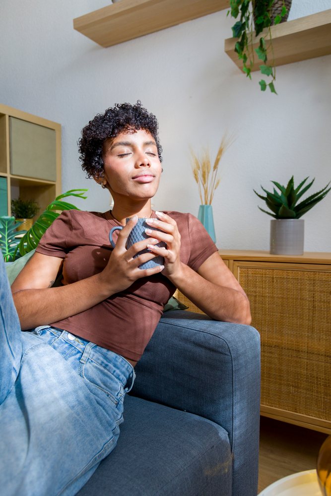 Young woman resting on a dark gray sofa, holding a ceramic coffee mug in a modern living room with indoor plants and natural sunlight.