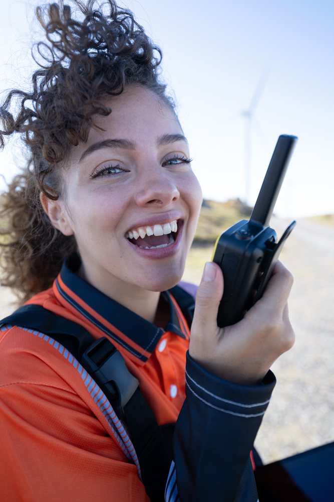 Smiling woman wearing a safety vest holds a walkie talkie outdoors near a wind turbine, sustainability, renewable energy, Galicia, Spain.
