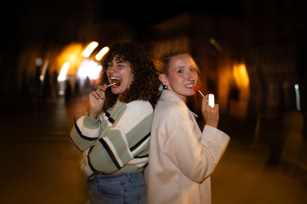 Two women smiling back to back while holding and eating lollipops on a street at night, friendship, Santiago De Compostela, Spain.
