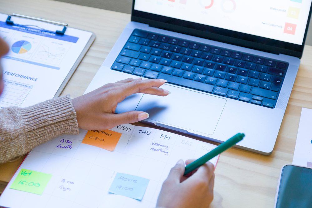 Person uses a laptop keyboard and writes on a calendar with sticky notes beside a business report in a home office setup, remote work.