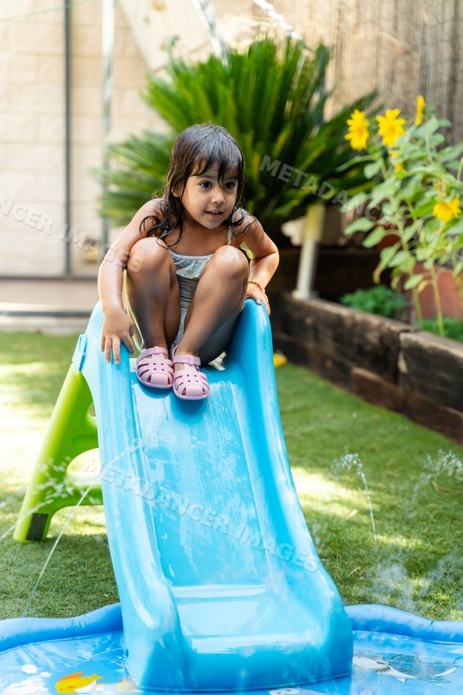 A little white girl sitting on a blue water slide in a backyard garden