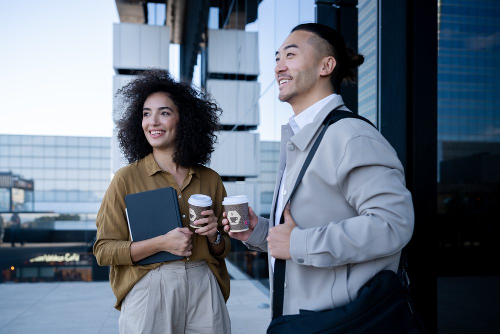 Two office workers smiling and talking while holding takeaway coffee cups on an outdoor terrace in a modern office setting, Madrid, Spain.
