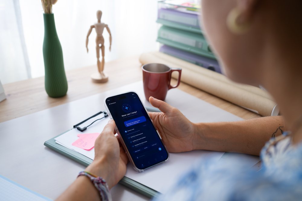 Person holding smartphone displaying an AI assistant app at a home office desk with office supplies, telecommuting, Spain.