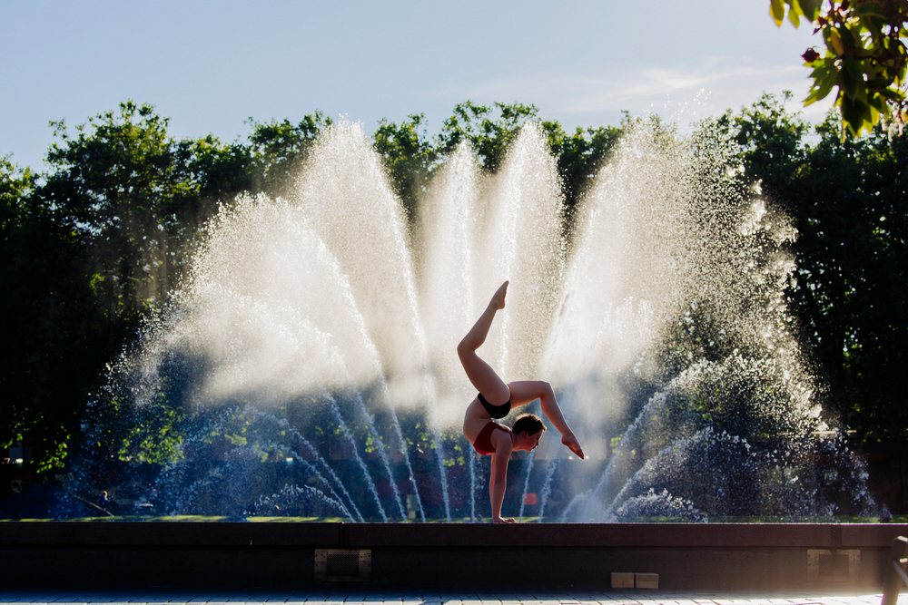 Acrobat performs a handstand balance pose in front of a water fountain with trees in an outdoor park, USA.