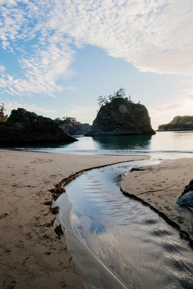 Stream flowing into the ocean on a sandy beach with rock formations under a cloudy sky, Boardman State Park, Oregon, USA.