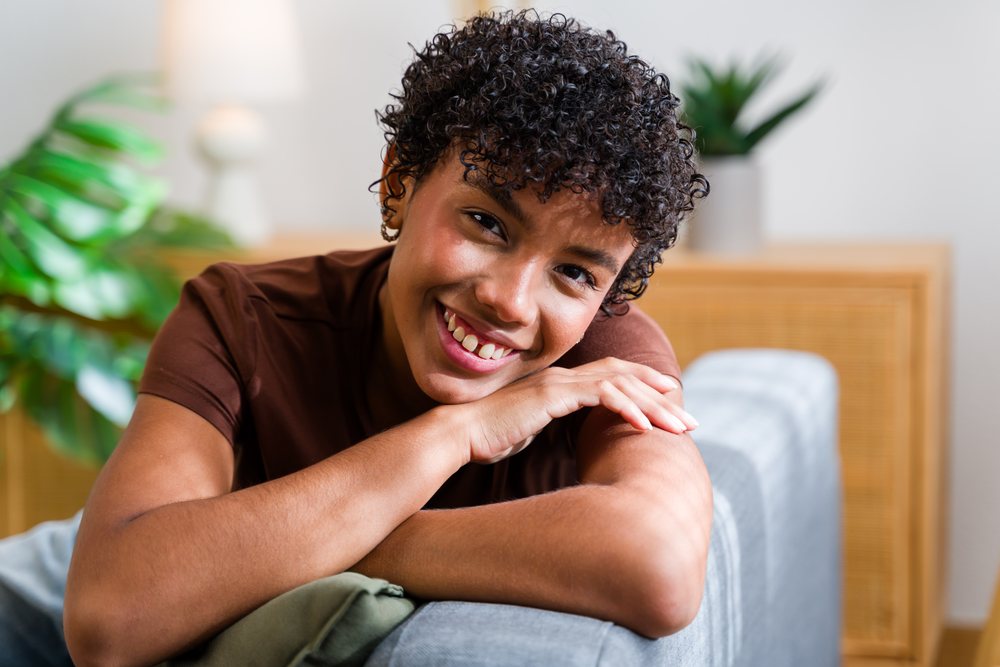 Smiling woman with curly hair wearing a brown shirt leans on the armrest of a gray fabric couch in a modern living room with indoor plants and natural light, Spain.