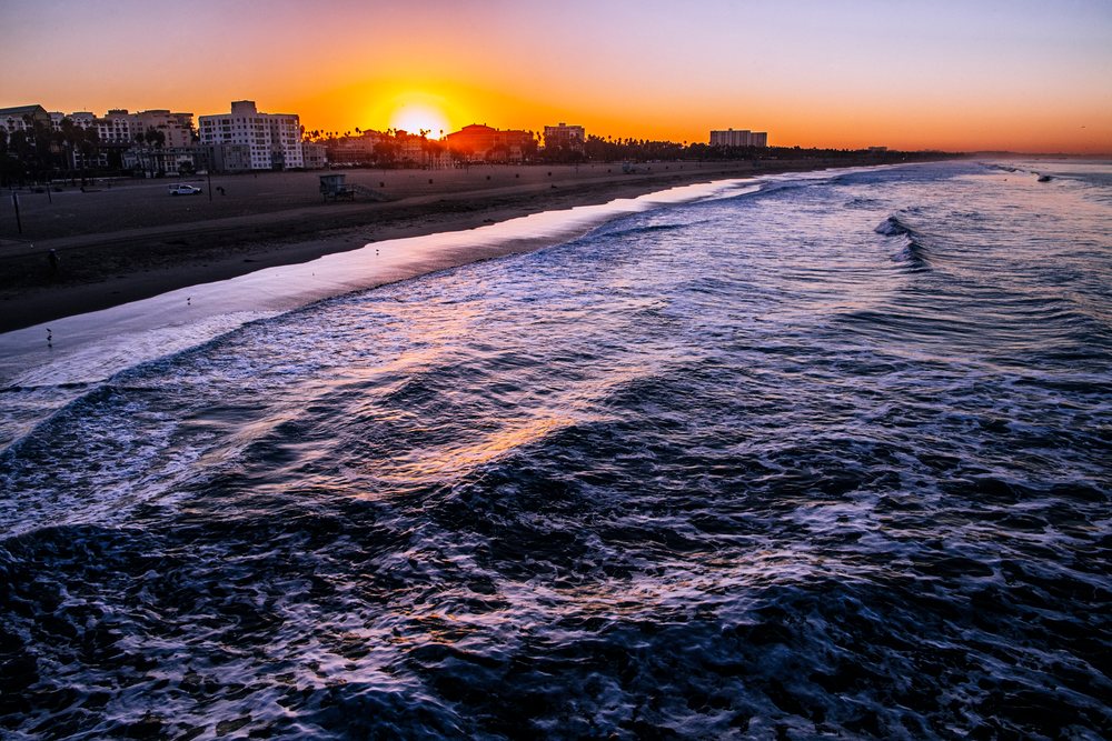 Sun sets over ocean waves rolling toward a sandy beach with buildings along the Santa Monica coastline in lifornia, CA, USA.