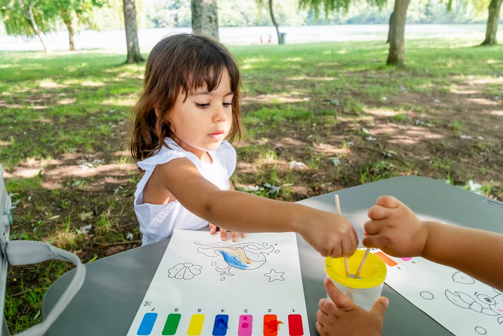 Children painting a bird coloring page with watercolors at a table outdoors in a grassy park by a lake, kids, Spain.
