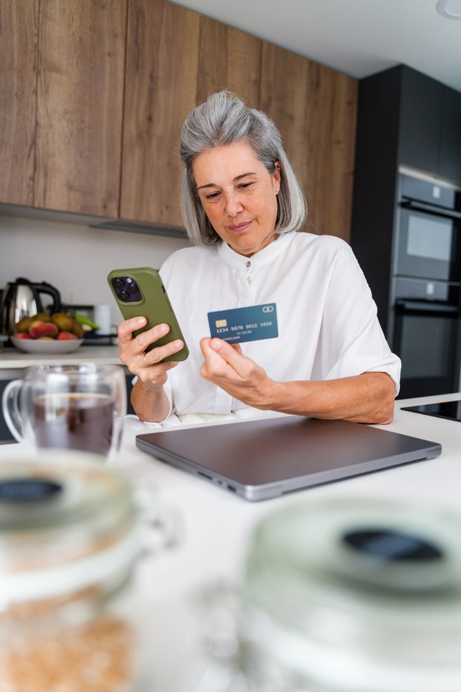 Woman checking a credit card while using a smartphone for an online payment at home kitchen, Spain.