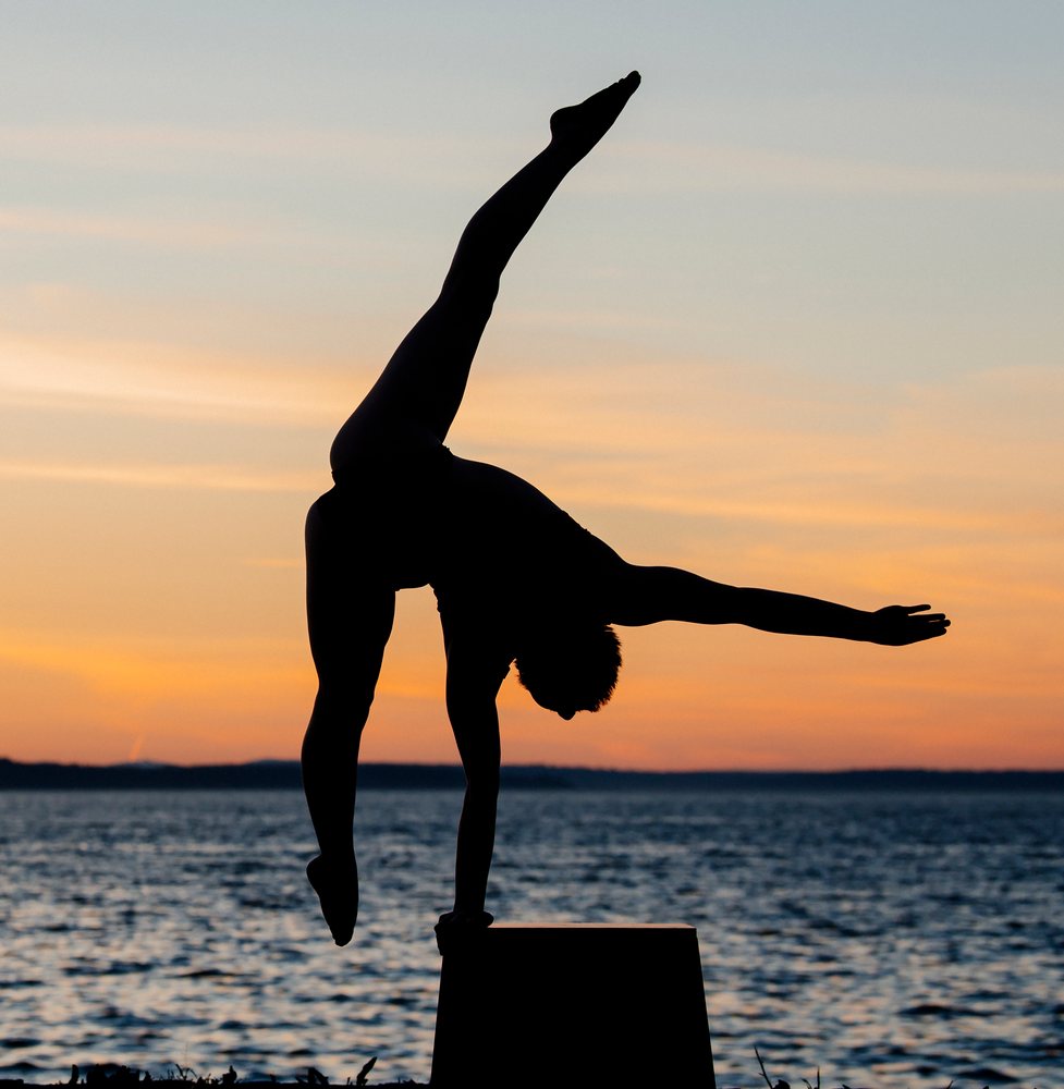 Silhouette of person performing a handstand on a raised platform by a lake at sunset with calm water and serene sky, USA.