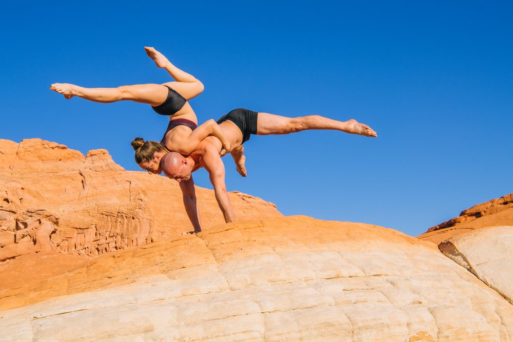 Two people perform an acrobatic yoga balance pose on a rocky desert landscape under a clear blue sky, USA.