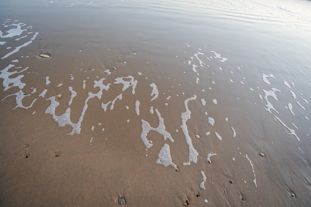 White sea foam and wet sand with small pebbles along a beach shoreline, kids, Spain.