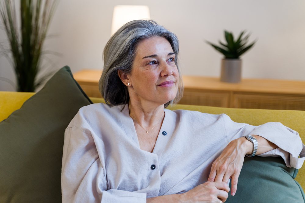 Thoughtful middle-aged woman with gray hair sits on a yellow sofa in a living room decorated with plants and a lamp, Spain.
