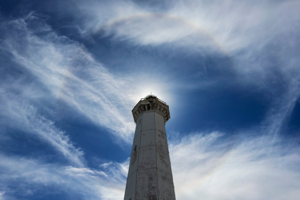 White Lighthouse tower with a sunbow halo glowing around its top under a cloudy blue sky, Torre Canne, Puglia, Italy.