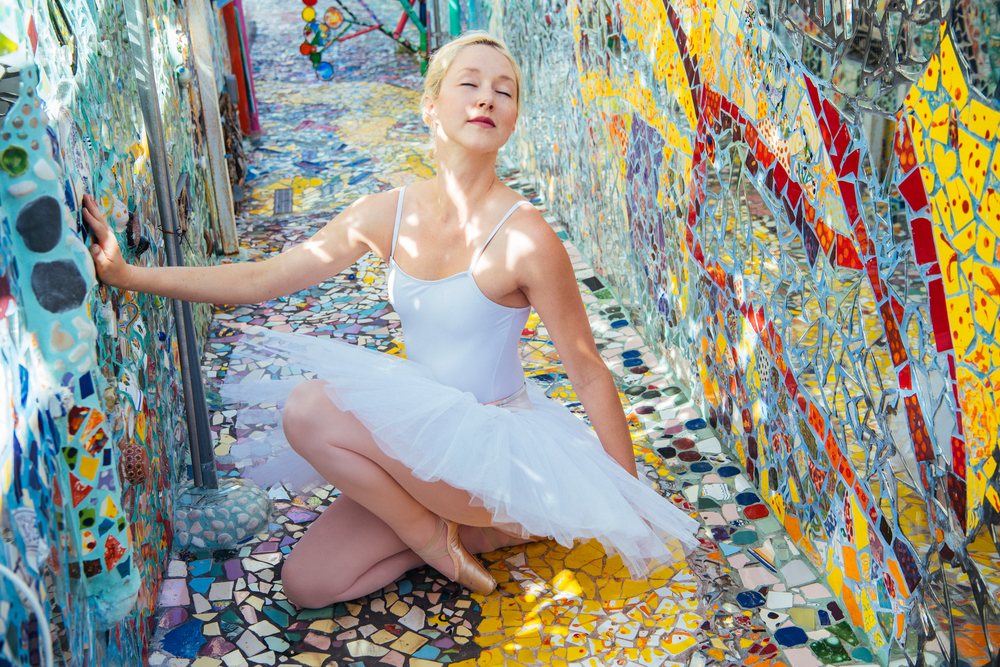 Female ballet dancer kneeling in white tutu and pointe shoes on colorful mosaic floor between mosaic walls in an outdoor California art installation, USA.