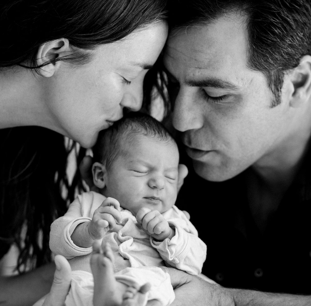 Newborn baby sleeping peacefully while being lovingly held and kissed by parents in an intimate black and white portrait, USA.