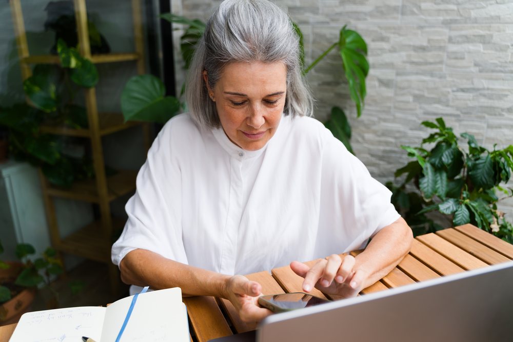 Woman with gray hair using a smartphone at a wooden table with an open notebook and indoor plants, Spain.