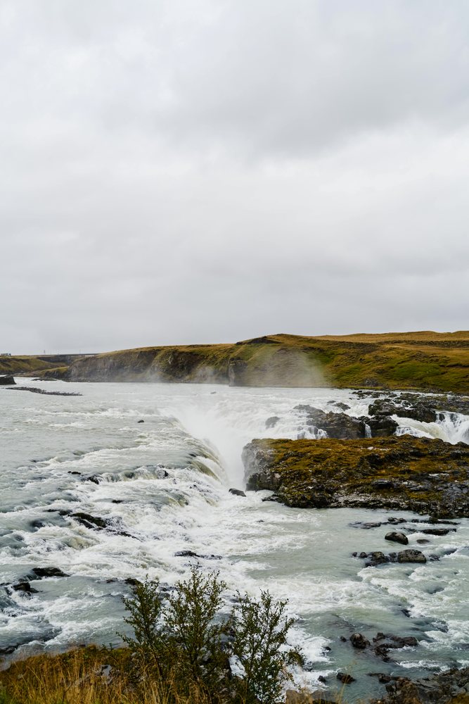 A powerful waterfall cascades over rocky cliffs into a river surrounded by grassy hills in Iceland under an overcast sky.