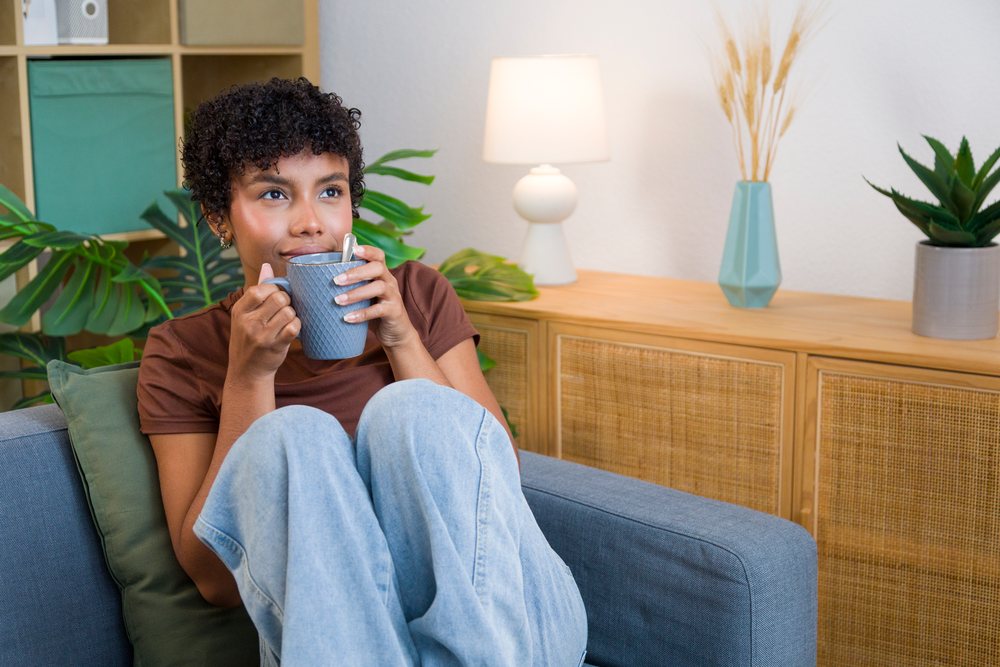 Woman seated on a sofa drinking a hot beverage from a gray mug in a cozy living room with plants and modern decor, Young woman, home.