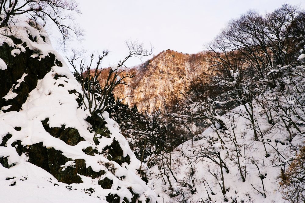 Sunlight illuminates a snow-covered mountain peak with bare trees and rocks on slopes, Nagano, Japan.