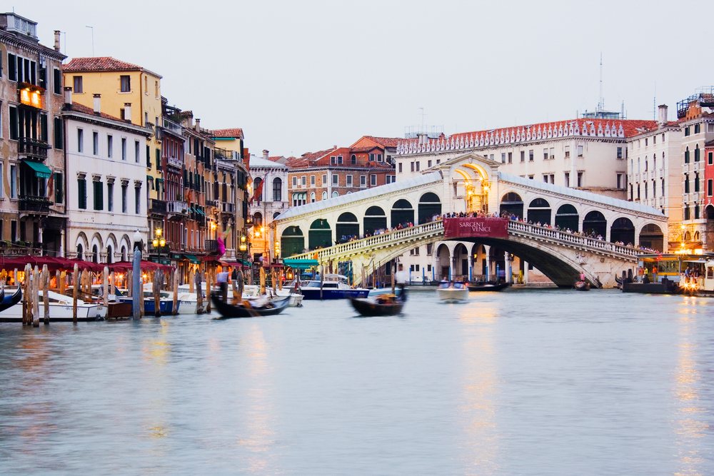 The Rialto Bridge spans Venice's Grand Canal with gondolas and boats moving on calm water reflecting historic buildings and lights, Veneto, Italy.