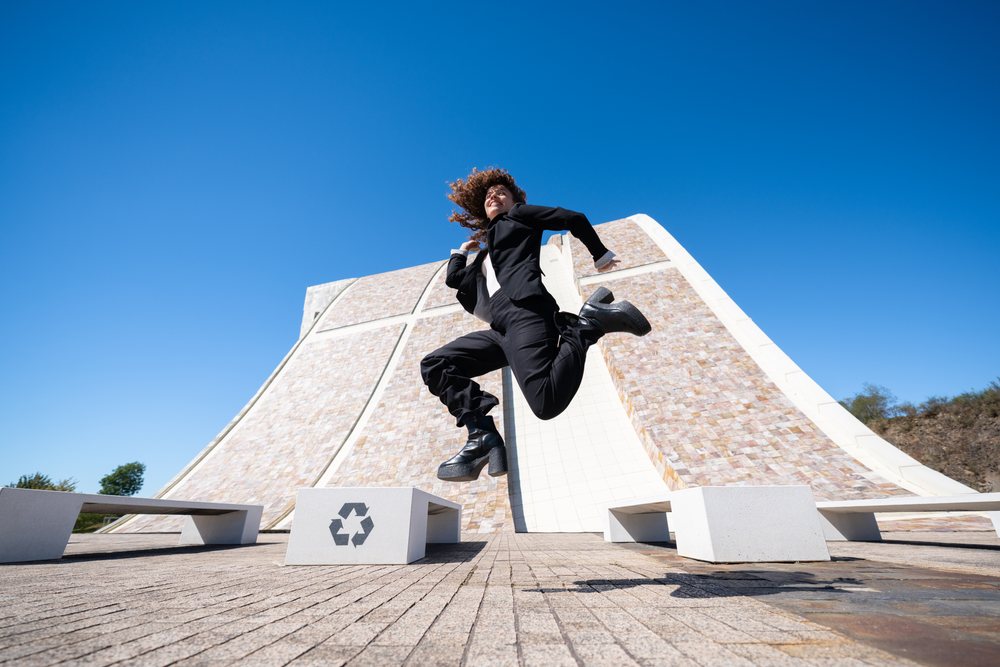 Professional woman jumping outdoors near benches with a recycling symbol in front of a modern curved building under a clear blue sky in Santiago de Compostela, businesswoman, traveling executive, Spain, Antiago De Compostela.