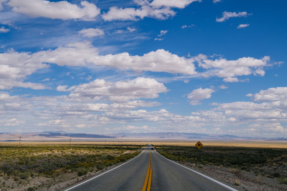 Open paved two-lane highway with yellow center lines stretches across desert terrain toward distant mountains under blue sky and white clouds, Nevada, USA.
