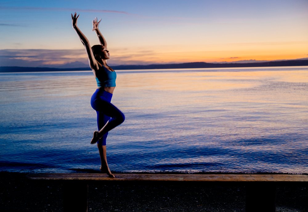Woman practicing a yoga balance pose on a bench by a lake at sunset, USA.