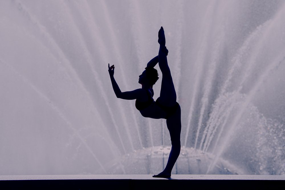 Silhouette of a woman performing a yoga dancer pose by a fountain with water jets outdoors, USA.