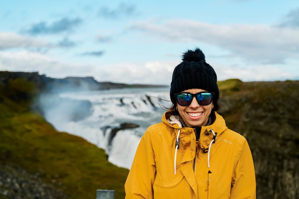A smiling woman wearing a yellow jacket and black knit hat stands near a waterfall, Iceland.