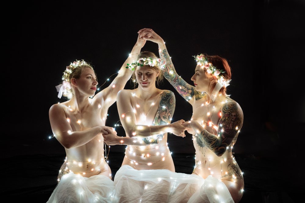 Three tattooed women wearing flower crowns, wrapped in fairy lights and white fabric, hold hands in a dark studio setting, muses, USA.