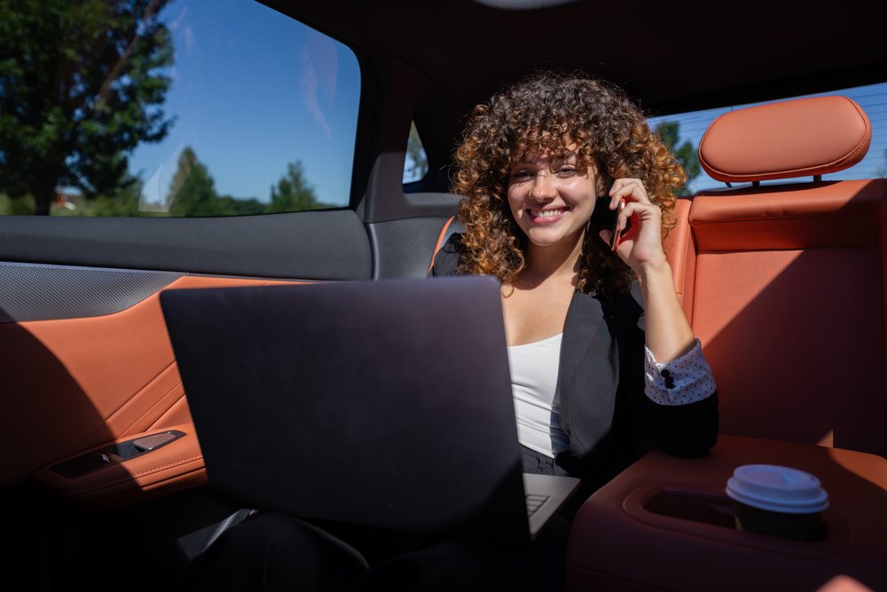 Woman with curly hair smiles while talking on the phone and using a laptop inside a car with orange leather seats and a coffee cup nearby, remote work, businesswoman, Spain.