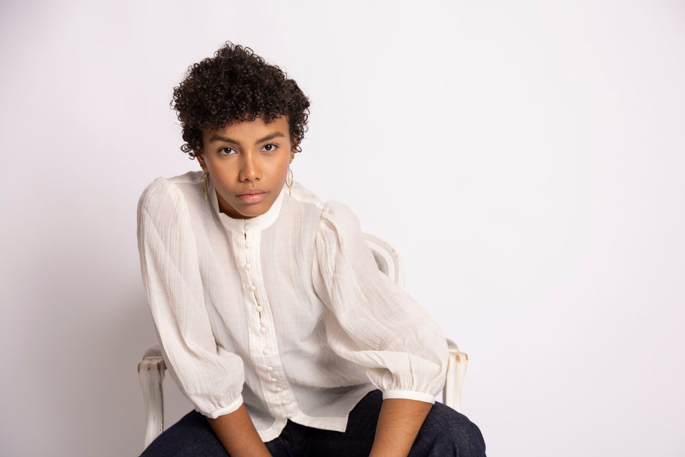 Young woman with curly hair wearing a white button-up blouse seated indoors against a plain backdrop, fashion, young culture.