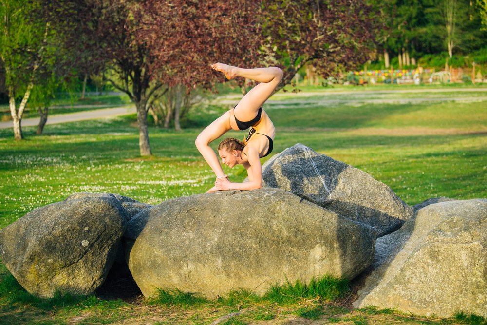 Girl wearing black bikini performing yoga stretch balancing on a large gray rock outdoors in a park with grass, flowers, and trees under sunlight, USA.