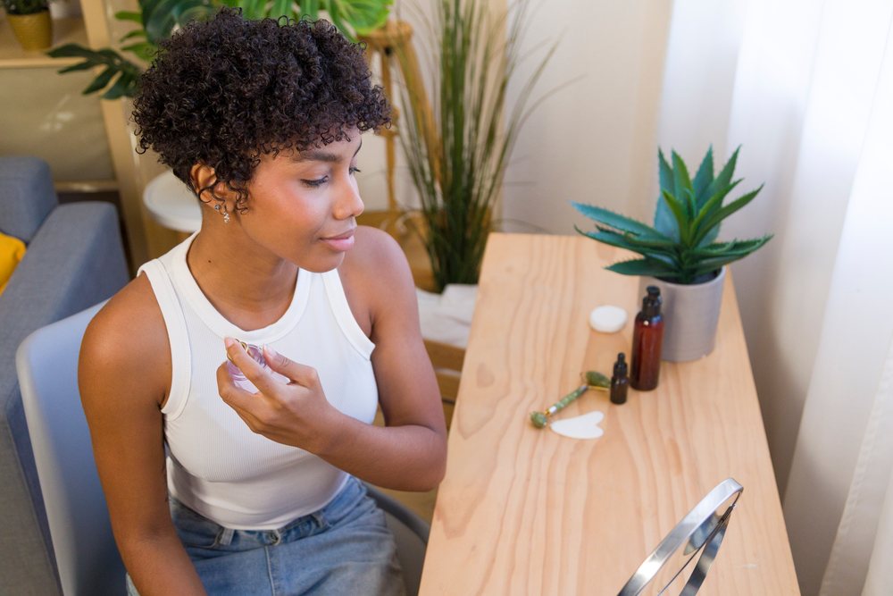 Woman with short curly hair applies perfume at a wooden table with skincare tools and a green plant in natural home light.
