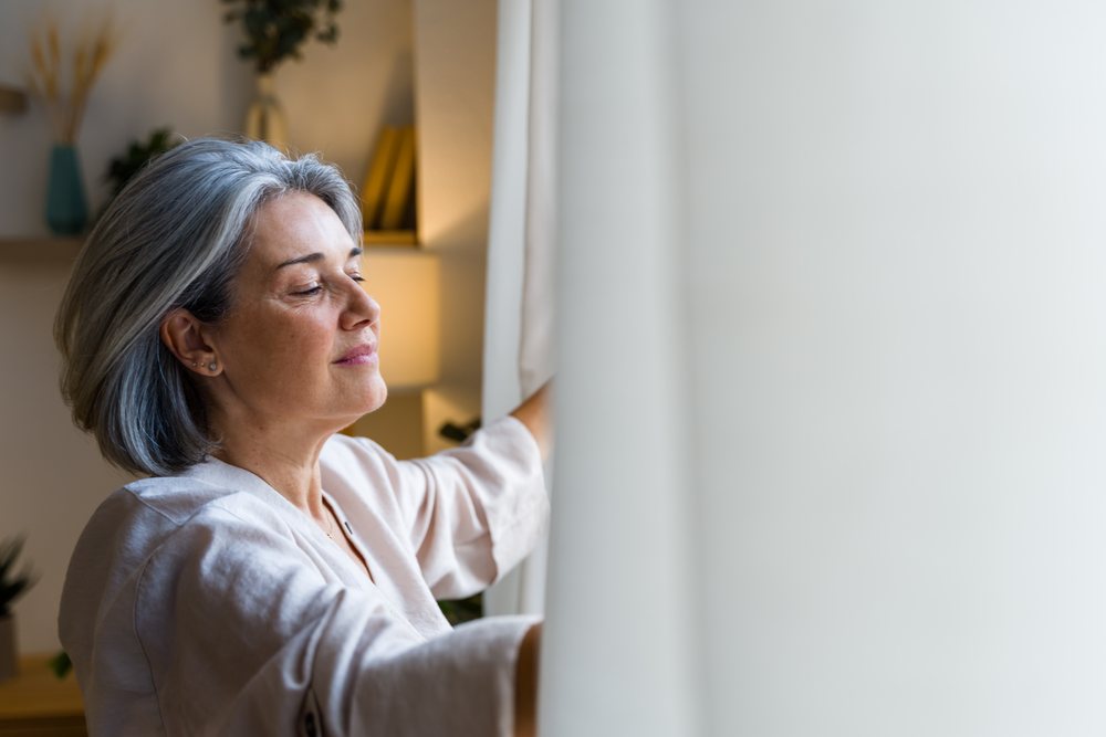 Woman with gray hair opening curtains in a peaceful indoor home setting, Spain.