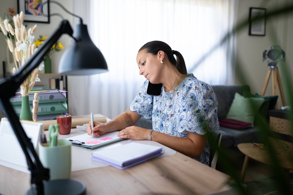 Woman multitasking by talking on phone and writing notes at a desk in a natural-lit home office, telecommuting, Spain.
