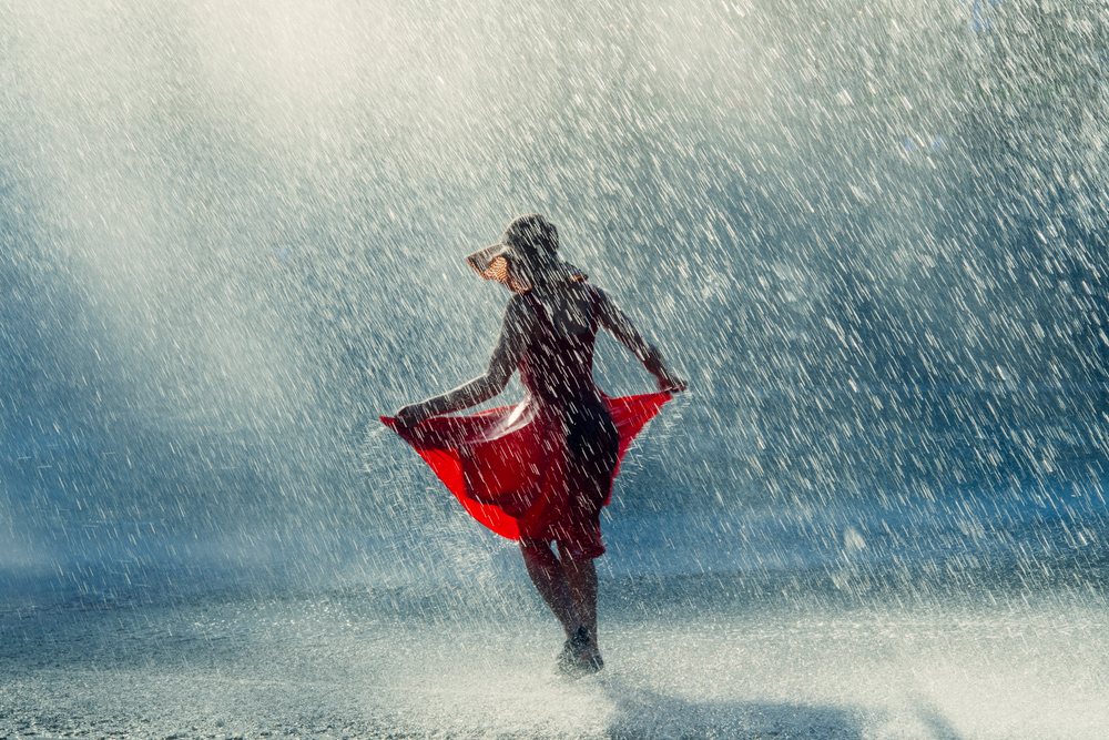 Woman dancing joyfully outdoors in rain wearing a red dress and sun hat with water droplets sparkling in sunlight, USA.