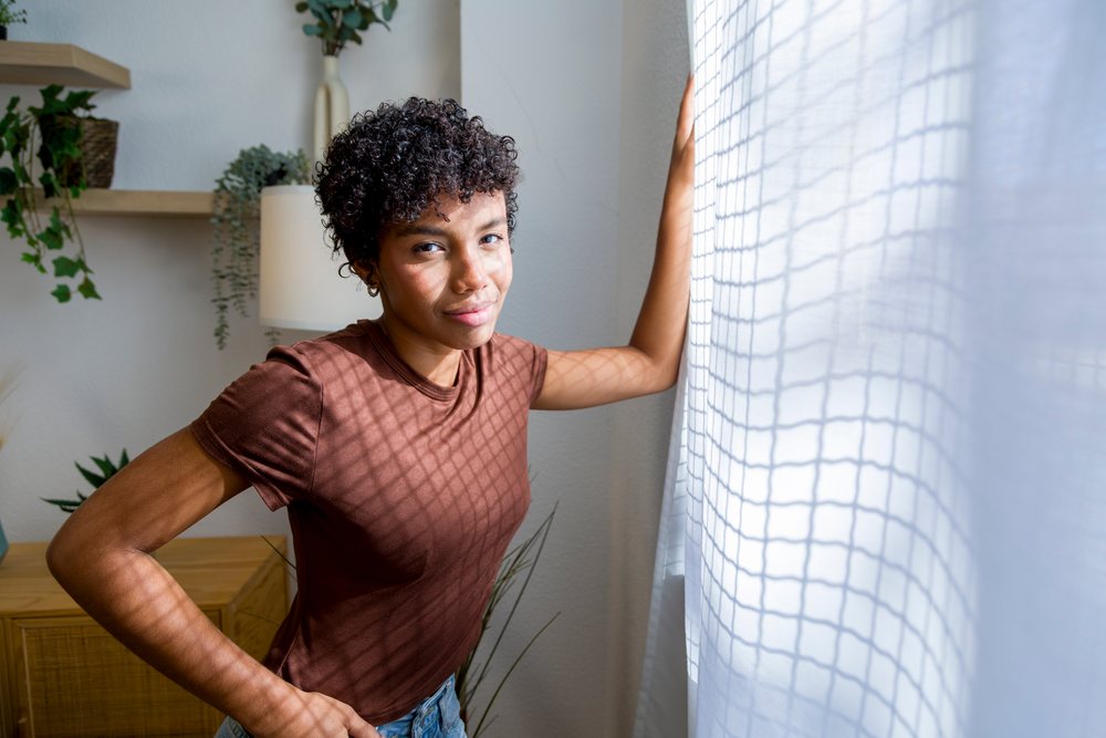 Young woman with curly hair in a brown shirt standing near a sheer curtain-covered window inside a home interior.