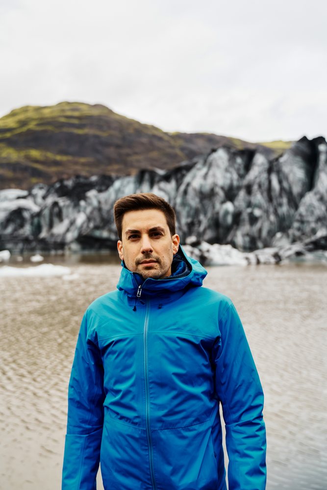A white man stands outdoors near a glacier and lake